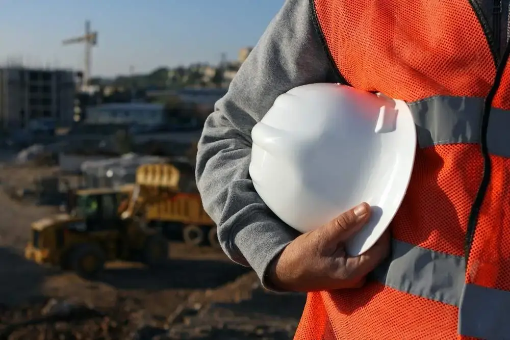 Construction worker wearing a high-visibility vest and holding a white hard hat, standing in front of heavy machinery on a construction site.