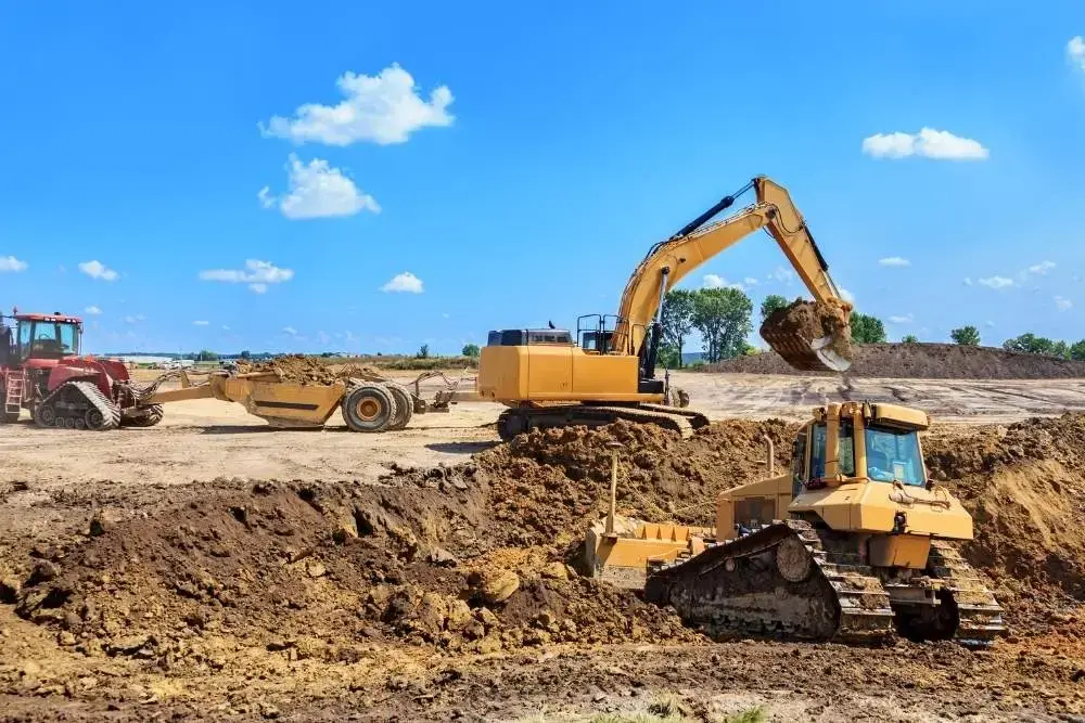 Earthmoving machines working on a wide dirt site under a blue sky. A large excavator lifts soil, a bulldozer pushes earth in the foreground, and a tractor with a scraper attachment sits in the background.