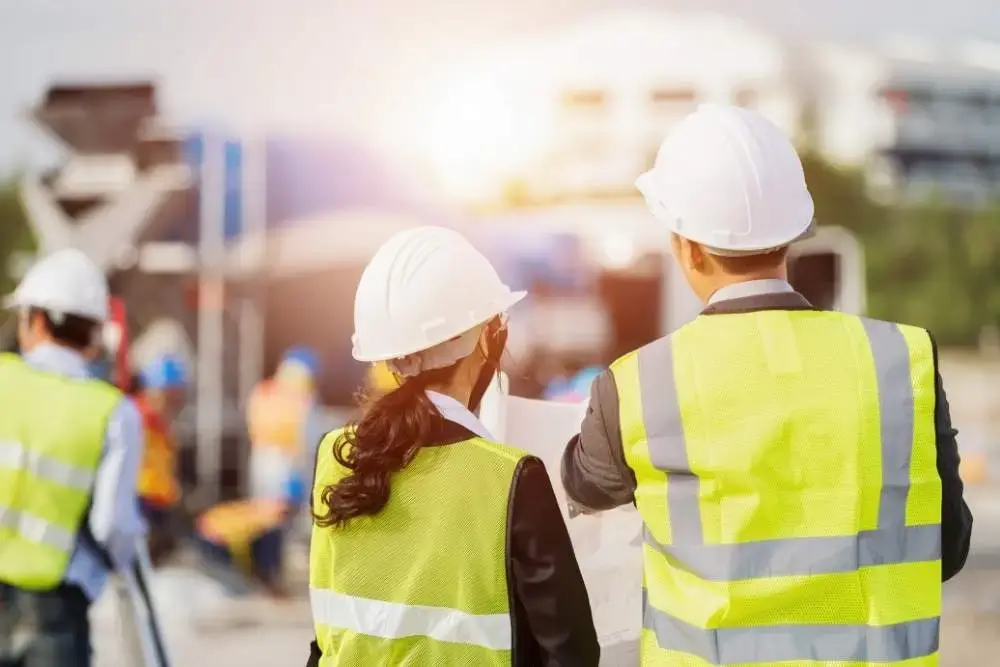 Two construction workers in white hard hats and high-vis vests review a site plan at an outdoor construction site, with other workers and machinery blurred in the background.